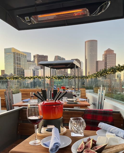a festive holiday setup on a rooftop bar, boston skyline in the distance, on the table is a red fondue pot and cocktails