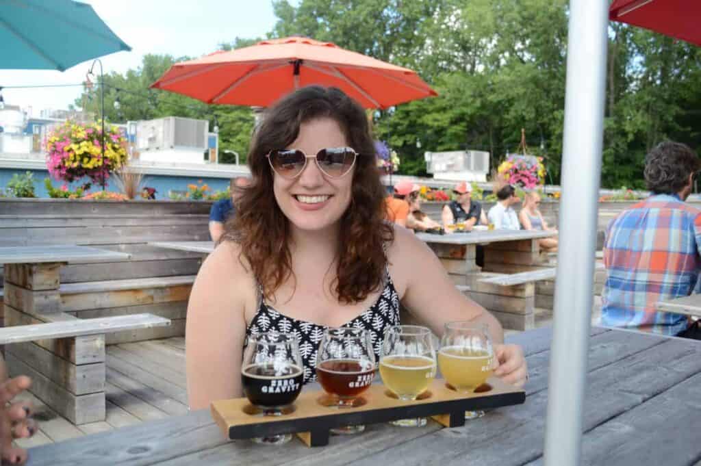 Smiling woman enjoying a beer tasting on a sunny patio in Vermont, with colorful umbrellas and a lively crowd in the background, suggesting a popular activity for visitors
