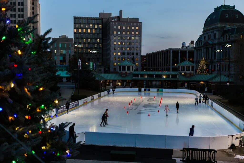 an outdoor skating rink in providence rhode island, city buildings seen in the background with a lit christmas tree close to the foreground