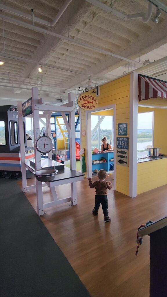 a toddler boy walks into a museum play area designed to look like a maine lobster shack