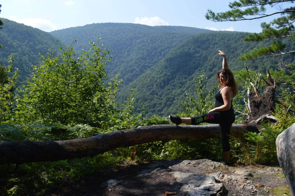 amy stands at a viewpoint on a hike stretching her leg like a ballerina