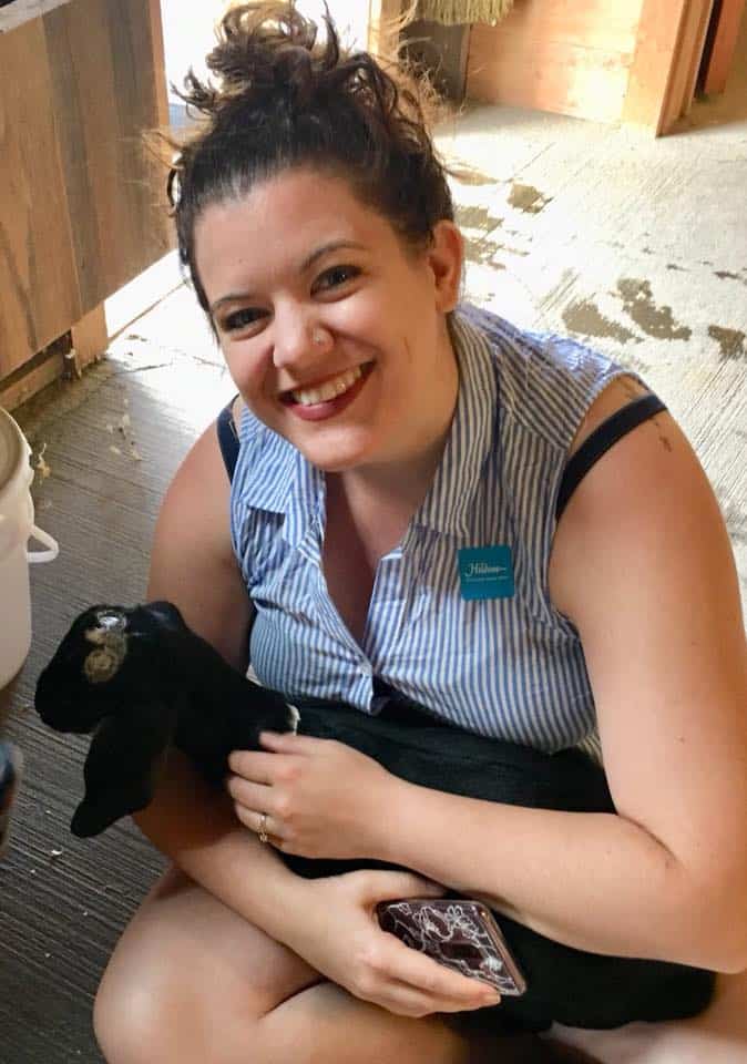 woman sits on the ground in a barn smiling and holding a small black goat