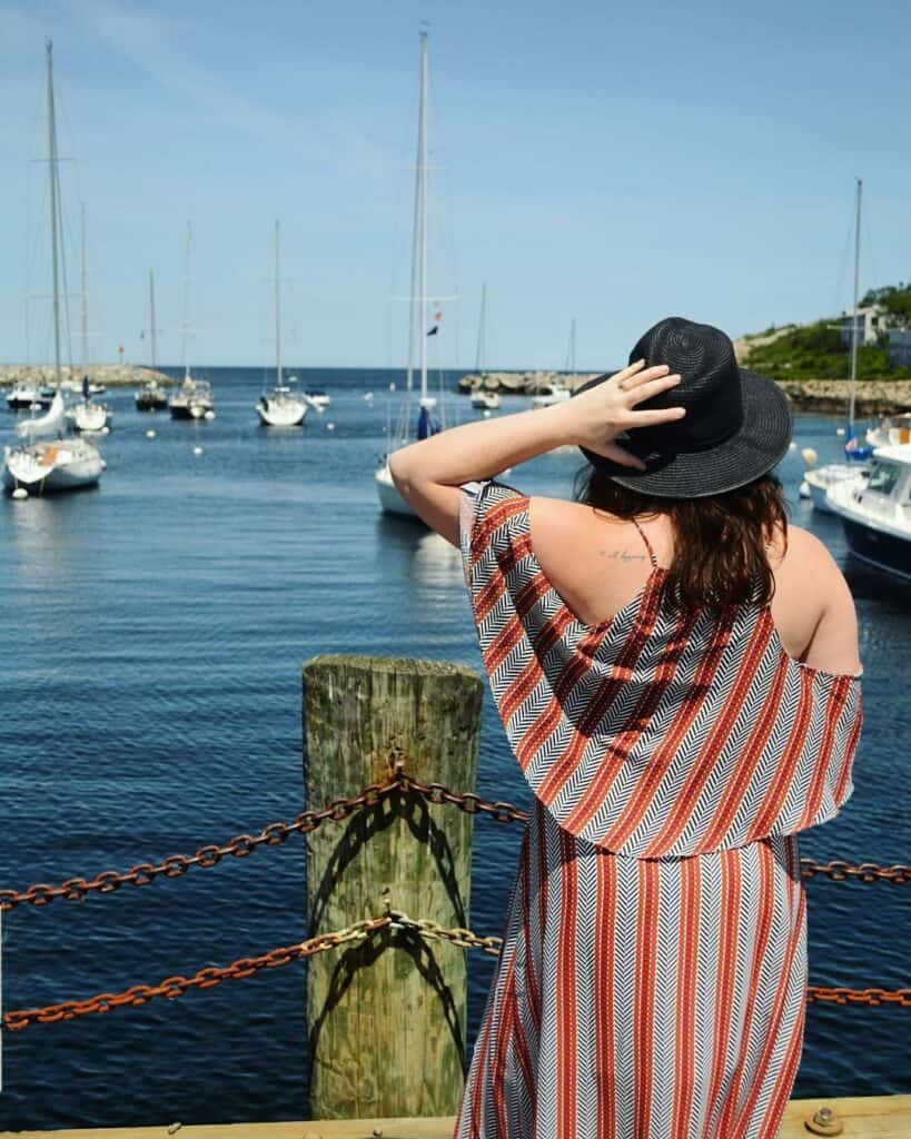 amy standing facing away from the camera with her hand on her black hat, boats in harbor in the distance