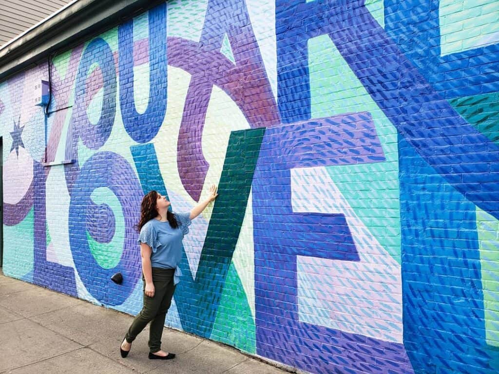 amy stands in front of a large wall mural that says you are loved