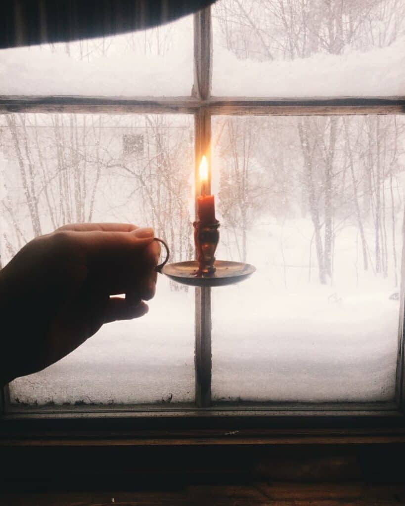 a frosted window looks out on a snowy scene, a hand holds a red candle in an old fashioned holder 