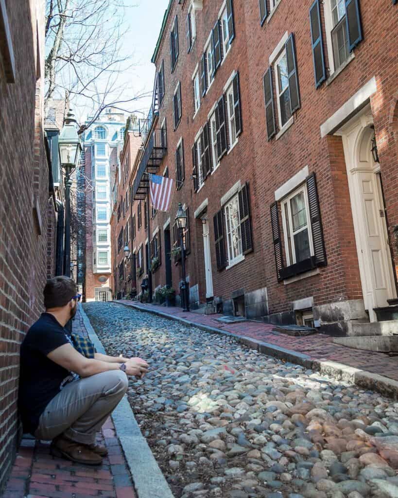 a man crouching against a brick wall on historic Acorn Street in Boston