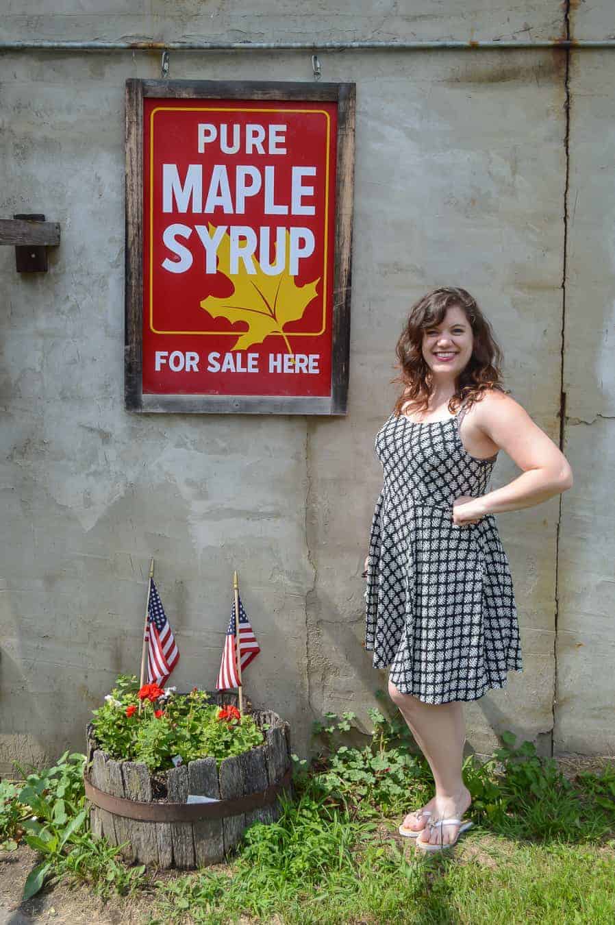 amy standing next to a sign that says pure maple syrup for sale here, she is wearing a blue dress and smiling, summertime in vermont