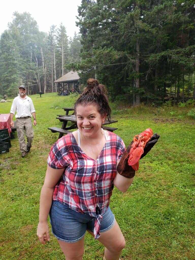 amy standing in a summer outfit holding a bright red fresh cooked lobster in her gloved hand. she is also soaking wet from a rainstorm