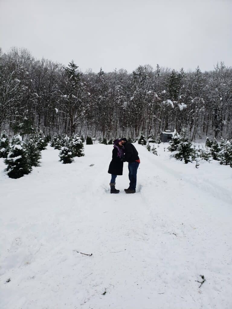 A couple stands in a snowy winter scene in Vermont, surrounded by tall pine trees at a christmas tree farm, sharing a Kiss on the cheek. 