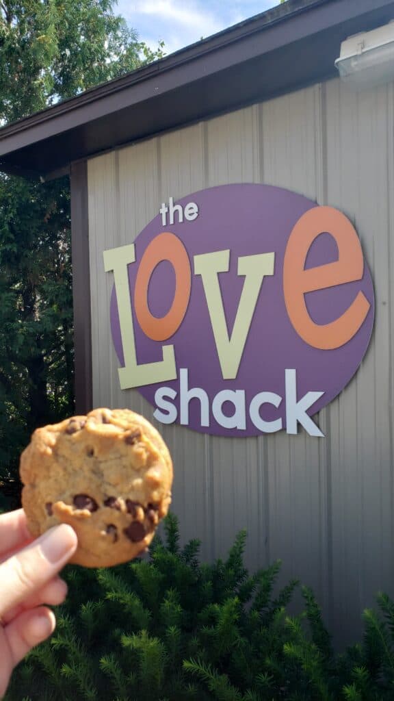Hand holding a freshly baked chocolate chip cookie in front of the sign for 'the LOVE shack', capturing a moment of sweet indulgence at a quaint dessert spot