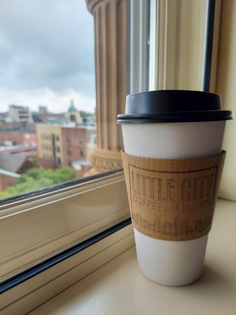 A takeout coffee cup from 'Little City Coffee' rests on a windowsill, with a view of the urban landscape and classical architecture in the background, evoking a cozy, reflective moment in a bustling city, Providence, Rhode Island