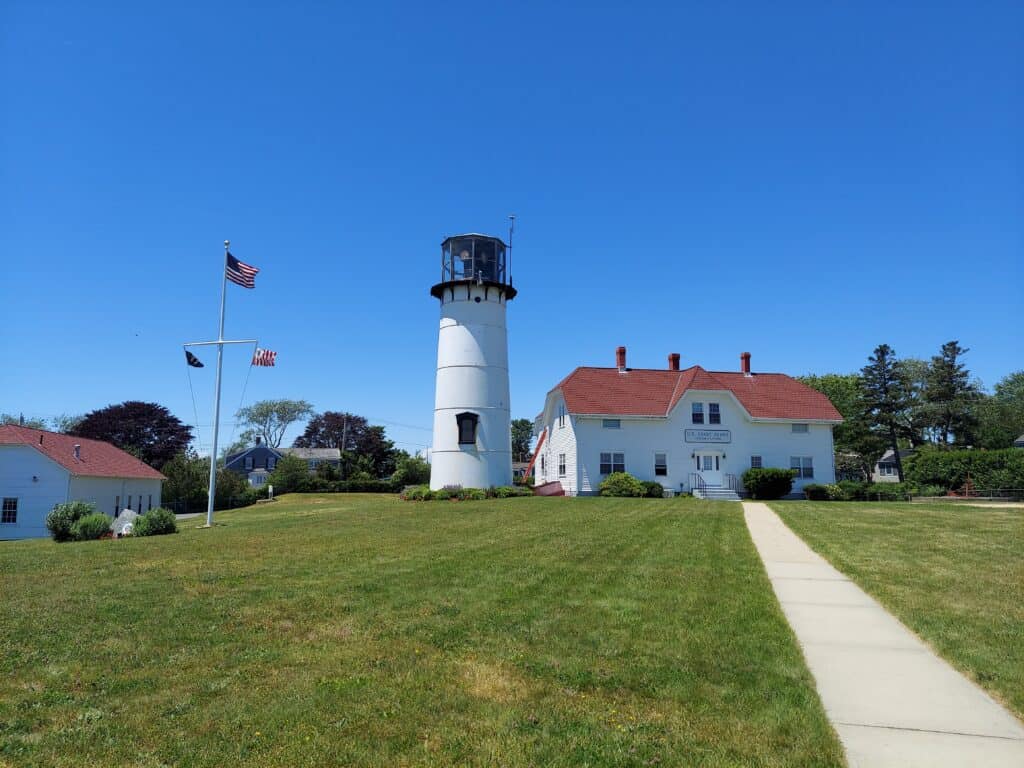 A classic New England lighthouse with a white facade and adjoining keeper's house, set against a clear blue sky. A well-tended lawn with a concrete pathway leads to the building, symbolizing the maritime heritage encountered while traveling through New England