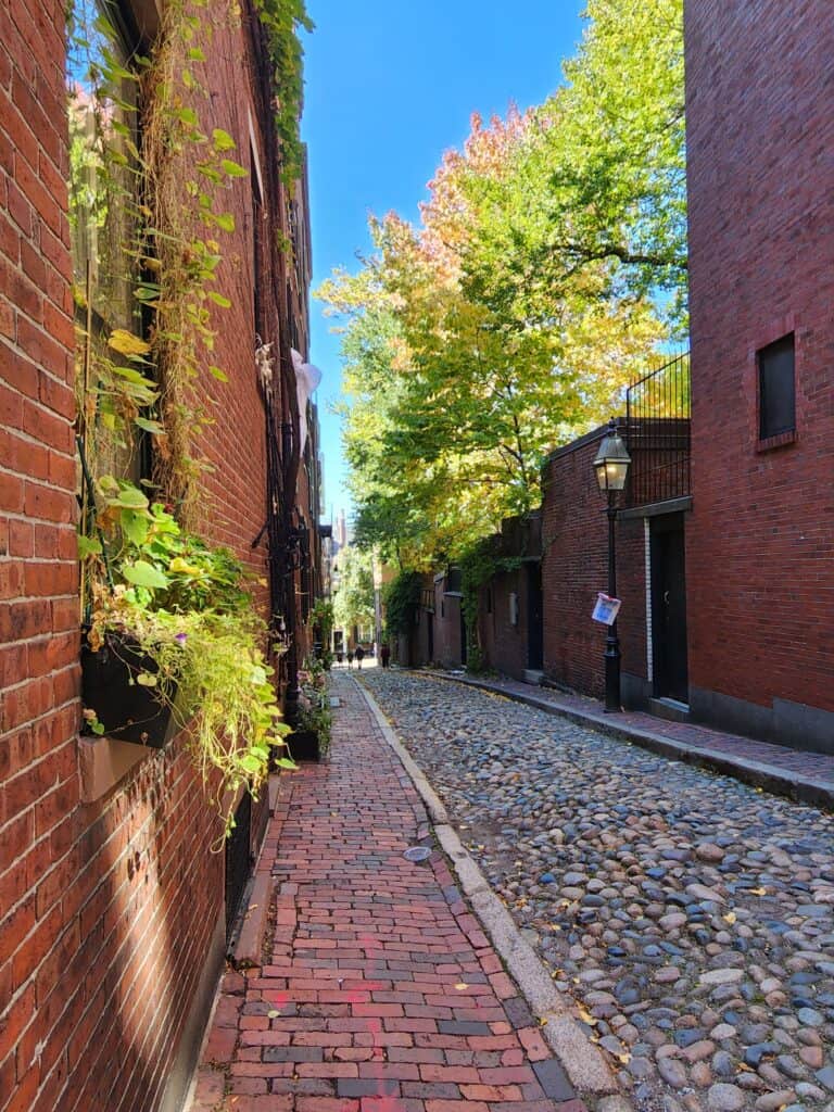 A quaint cobblestone alley in Boston, flanked by historic red brick buildings with greenery, capturing the essence of the charming places to visit in Boston