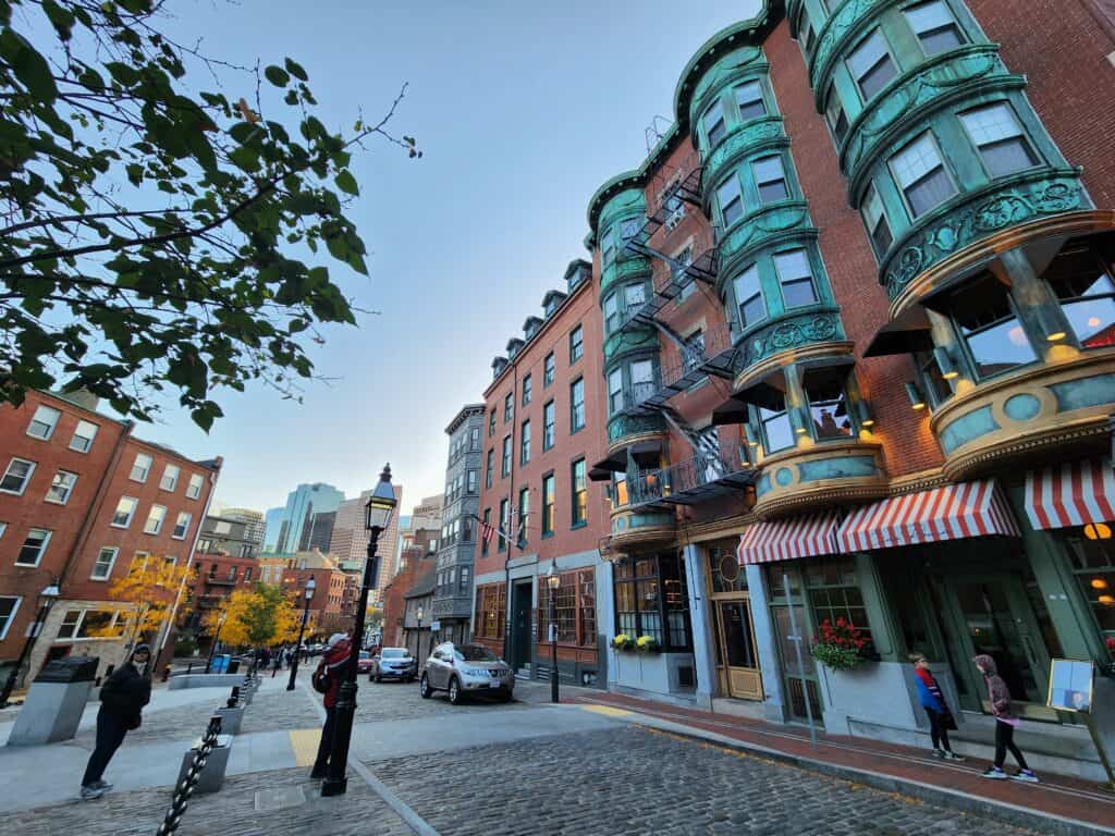 A street view in Boston, capturing the essence of a New England road trip itinerary, with cobblestone streets and historic brownstone buildings, featuring distinctive green bay windows. The scene is set against a backdrop of modern skyscrapers, merging the old with the new in this iconic cityscape.