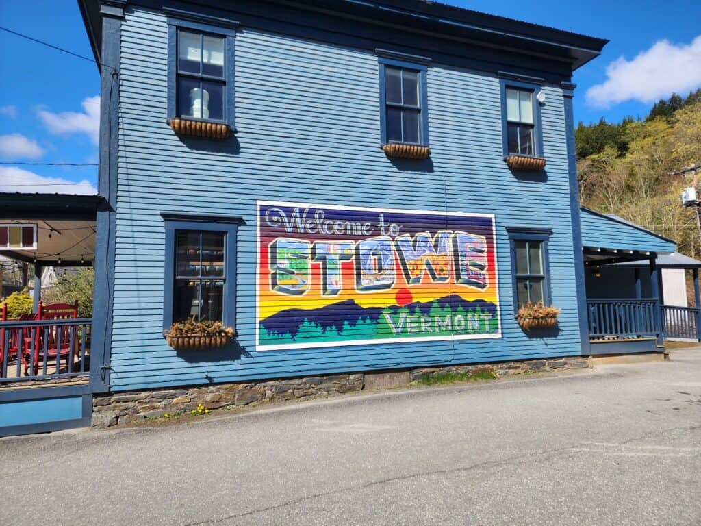 A vibrant 'Welcome to Stowe, Vermont' mural painted on the side of a classic blue New England building, complete with hanging flower baskets. This colorful greeting sets the tone for a picturesque stop on a road trip through New England.