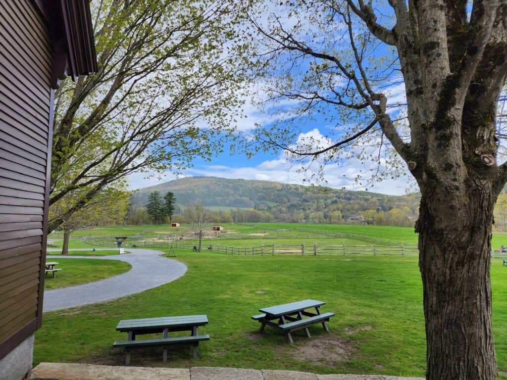 Picnic tables nestled near a winding pathway with a scenic view of the lush green fields and rolling hills in Vermont, offering a perfect spot for a leisurely outdoor meal in the countryside.