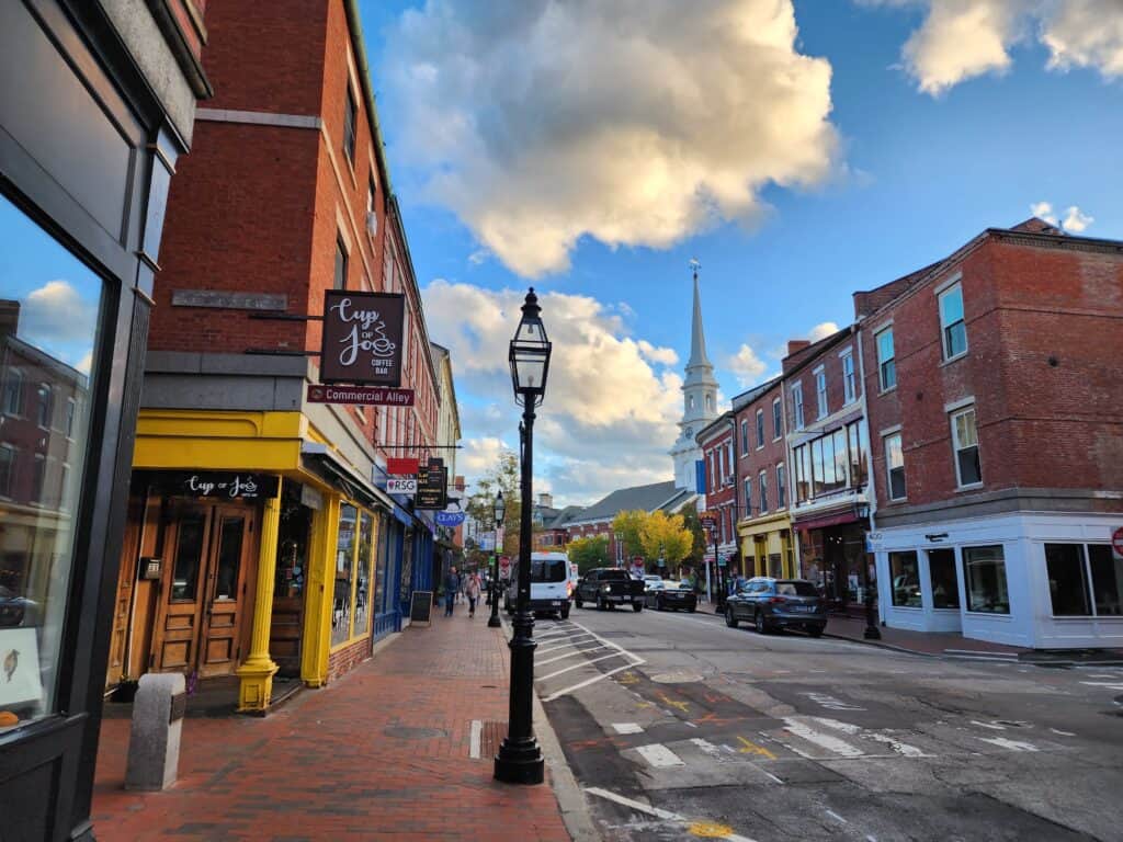 A charming street in a New England town during a getaway, featuring historic brick buildings with a quaint coffee shop, and the steeple of a white church rising against a sky with puffy clouds. The inviting atmosphere is perfect for a leisurely stroll and exploration.