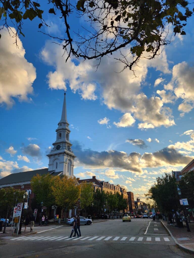 The historic charm of a New England street captured in the golden hour, featuring pedestrians crossing, a classic church spire rising into the sky, and the dynamic play of sunlight and clouds above.