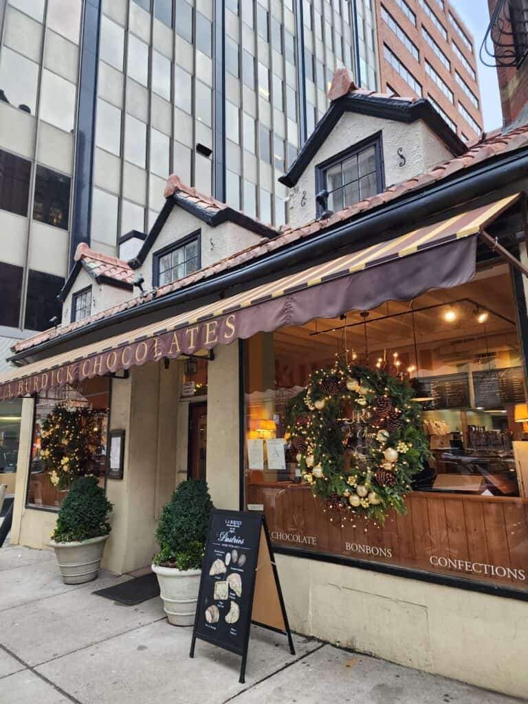 The charming façade of L.A. Burdick Chocolates, nestled in Boston, showcases festive holiday decorations with a large wreath and twinkling lights, inviting visitors to indulge in gourmet chocolates and confections