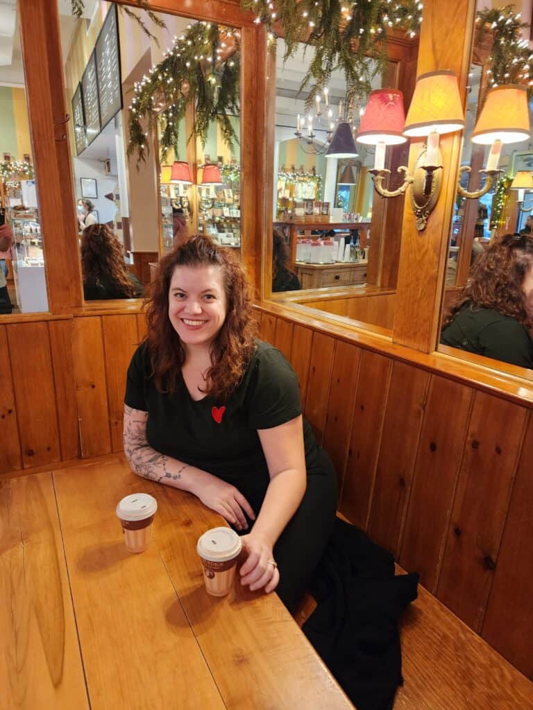 A woman sits at a wooden booth with two cups of coffee on the table, smiling in a warmly lit café adorned with festive decorations, reflecting a cozy and inviting atmosphere