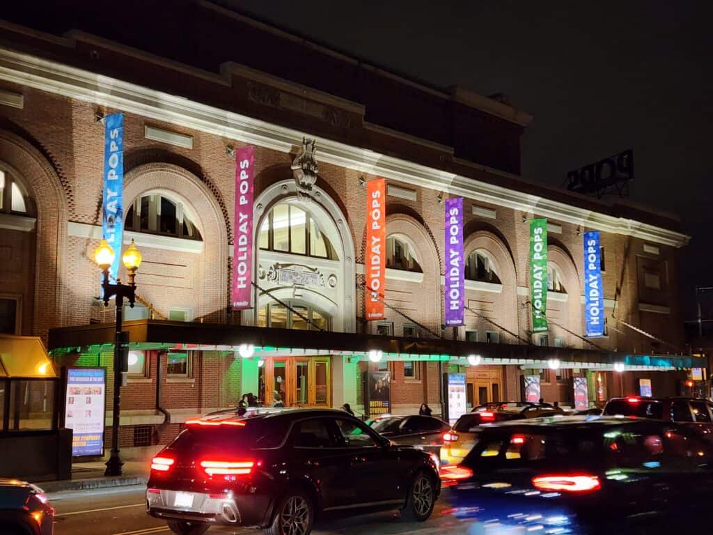 Outside of Boston symphony hall at night. Colorful signs hang from the building while cars drive by in front