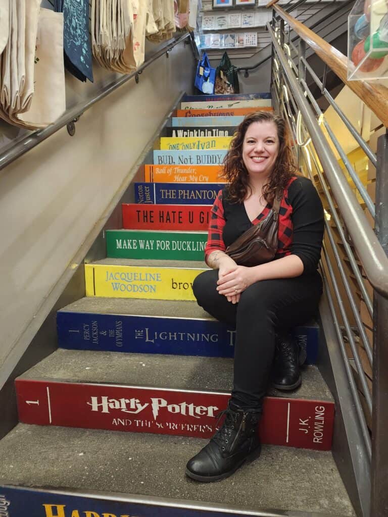 A woman sits smiling on the creatively painted literary staircase inside a Boston bookstore, each step resembling a book spine, a charming addition to Boston bucket list activities for book lovers