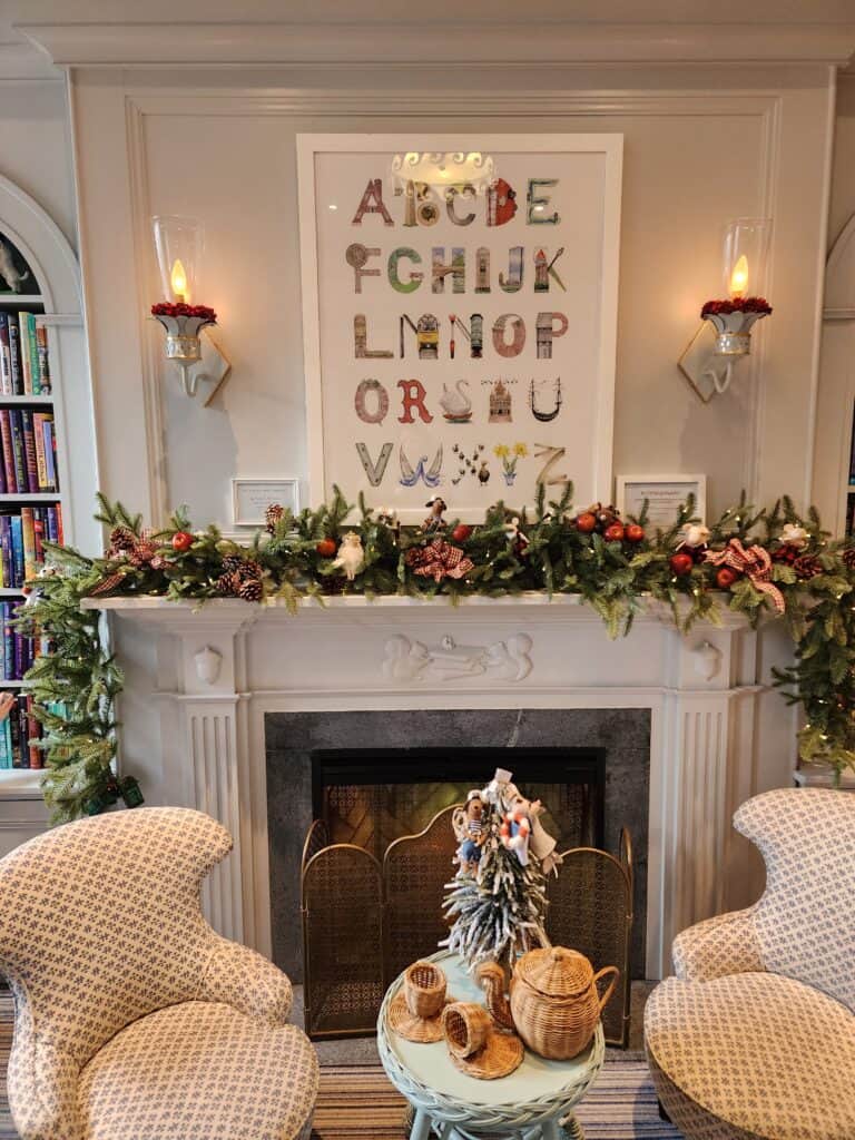 An elegant fireplace mantel is adorned with a lush holiday garland and flanked by two wall sconces, above which hangs a whimsical alphabet artwork, all set in a cozy room with patterned armchairs and a festive tabletop tree