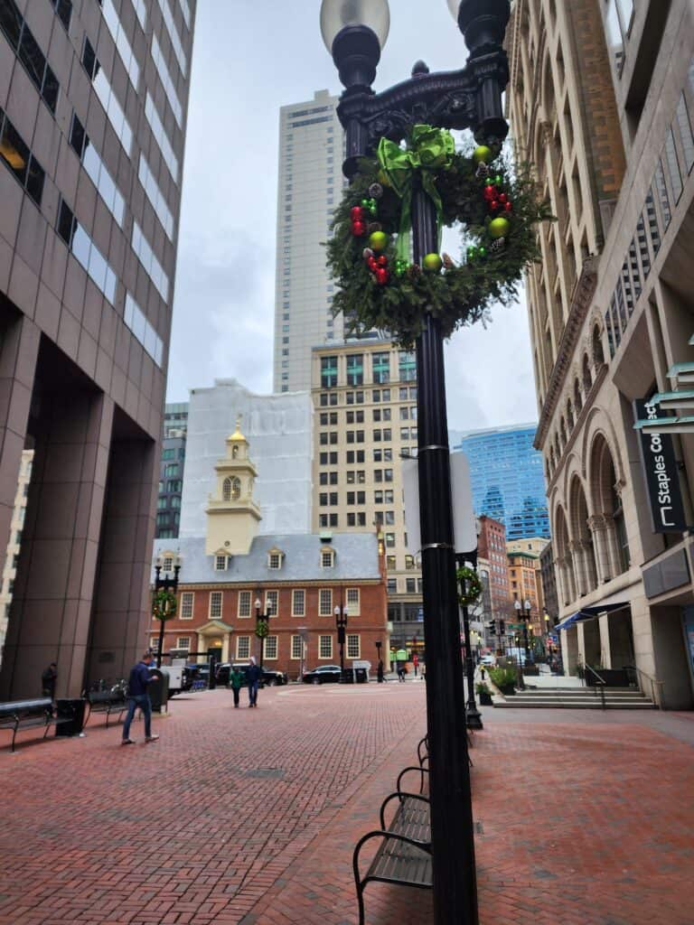 A wreath festooned with red and green ornaments hangs on a lamppost, framing a view of the historic Old State House in Boston, juxtaposed with modern buildings under an overcast sky, reflecting the city's blend of history and modernity