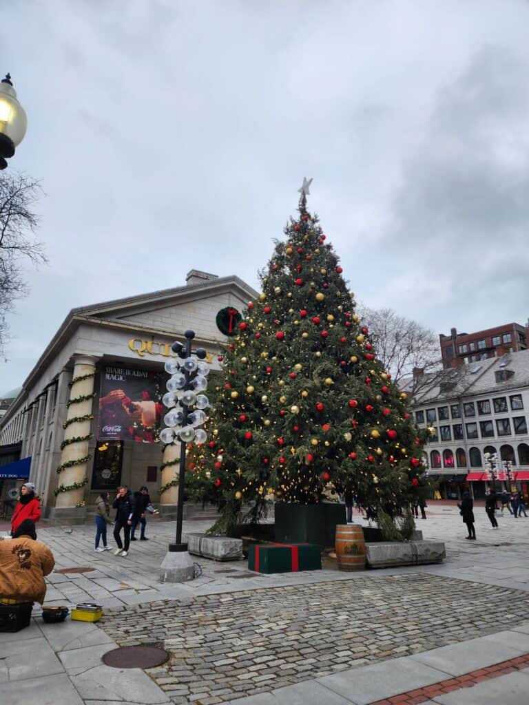 A majestic Christmas tree stands tall adorned with red and gold ornaments and a shining star topper, set against a backdrop of historic buildings and cobblestone streets in a Boston square, evoking a traditional holiday spirit