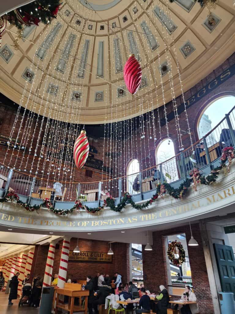 Inside Quincy Market, a bustling Boston marketplace, a grand dome ceiling is adorned with cascading fairy lights and oversized hanging Christmas ornaments, surrounded by balconies decked with garlands and wreaths, creating a festive holiday shopping atmosphere