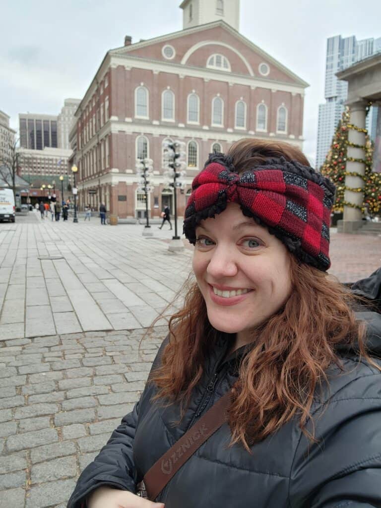Smiling woman in a plaid headband and winter jacket posing in front of Faneuil Hall, a must-visit location on any Boston bucket list, with festive Christmas decorations in the background