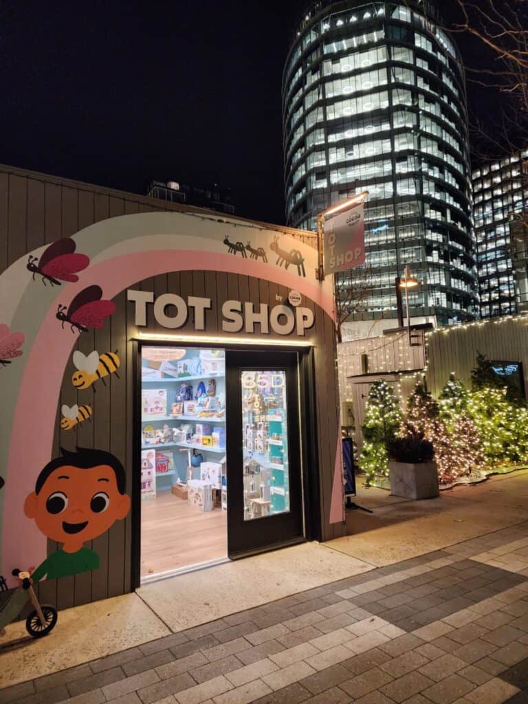 A small shop that is called Tots Shop sits open at night in a city with large skyscrapers behind
