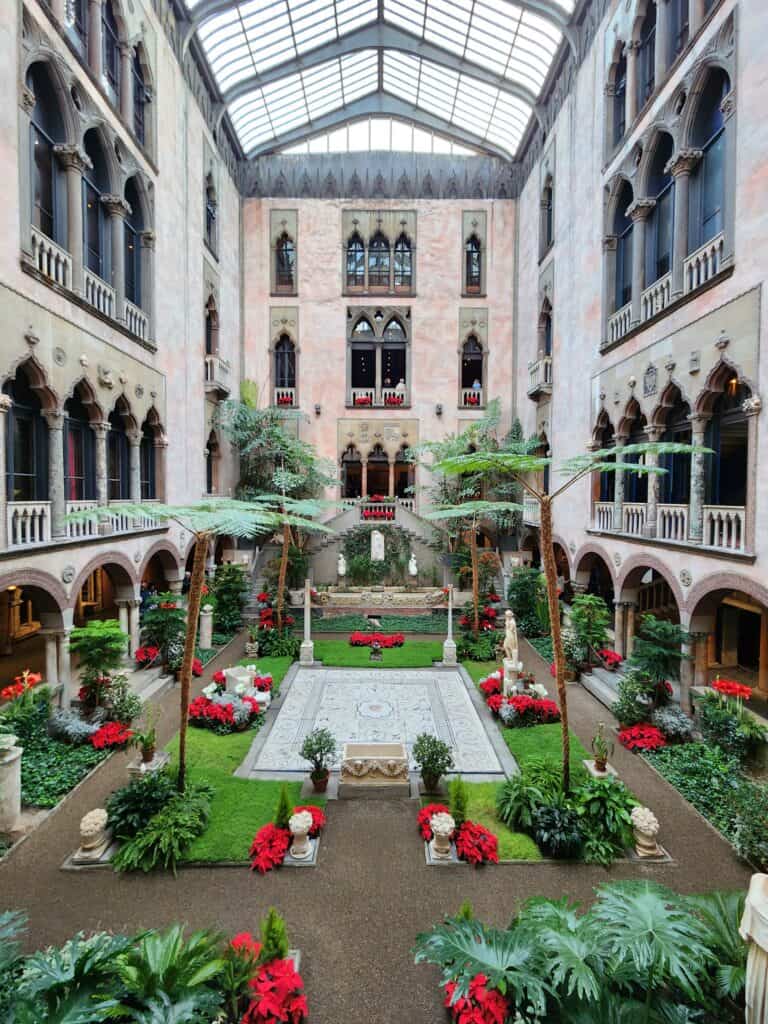 The interior courtyard of the Isabella Stewart Gardner Museum in Boston, adorned with vibrant red poinsettias and lush greenery, under an elegant glass ceiling, creating a serene and historically rich environment.