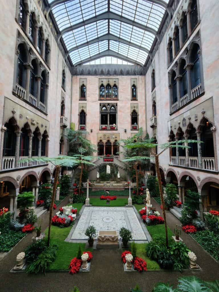 Lush courtyard garden inside the Isabella Stewart Gardner Museum in Boston, MA, showcasing a Venetian-style palazzo architecture with a glass roof, balconies, and vibrant red poinsettias.