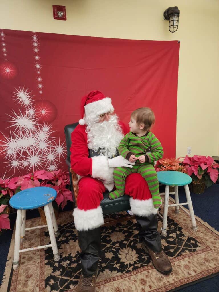 A child in festive green pajamas shares a moment with Santa Claus, seated together against a red backdrop with white snowflake decorations, conveying a heartwarming holiday scene