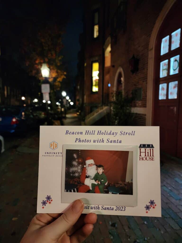 A hand holds a photograph from the Beacon Hill Holiday Stroll featuring a child sitting with Santa Claus, set against the backdrop of a dimly lit, historic Boston street at night, capturing a special moment from the event