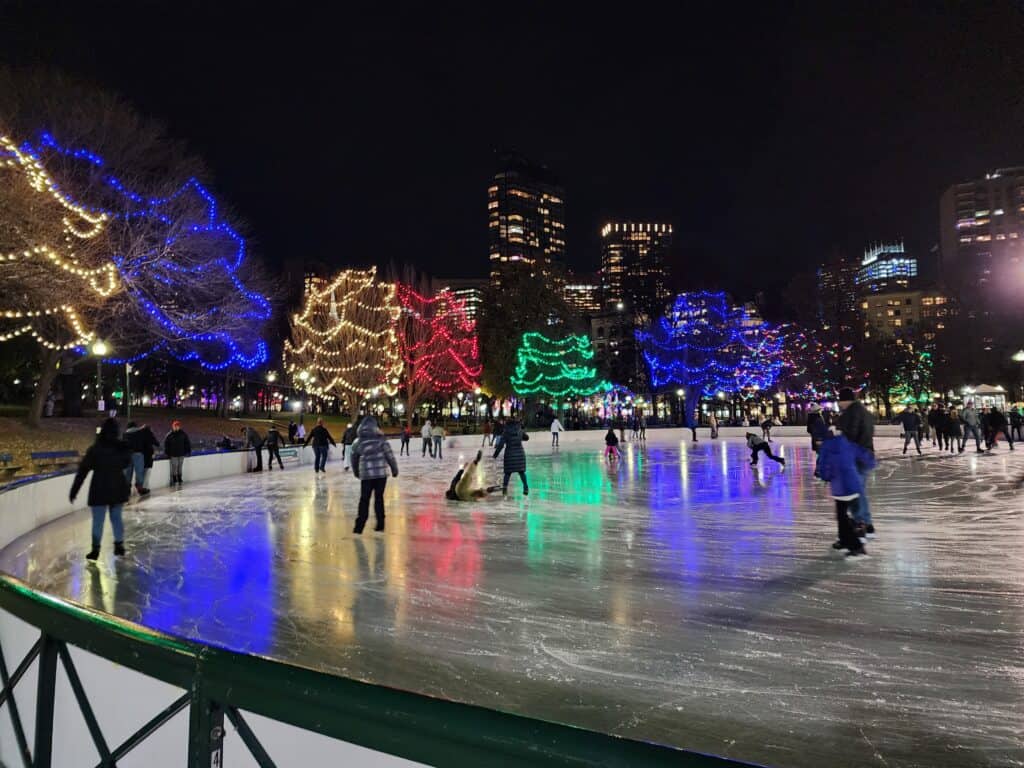 Skaters glide on an ice rink at night in Boston, under a canopy of trees adorned with twinkling lights in shades of blue, yellow, and red, with the city's glowing skyline in the background