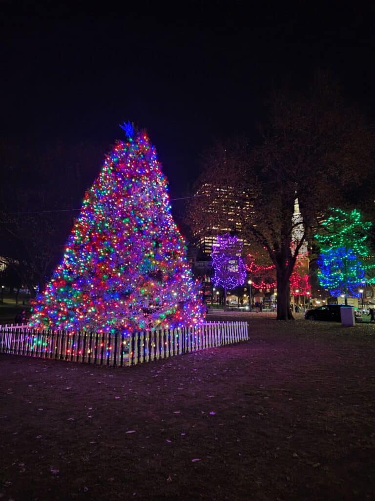 A vibrant Christmas tree adorned with colorful lights stands prominently in an evening park setting, surrounded by a white picket fence, with more illuminated trees in the background contributing to a festive atmosphere.