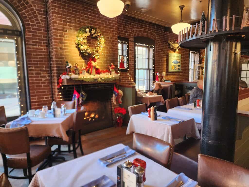 A cozy and inviting restaurant interior, with exposed brick walls and festive Christmas decorations. A beautifully adorned wreath hangs above the fireplace, adding a warm holiday ambiance to the dining area set with white linen tables, awaiting guests for a seasonal feast