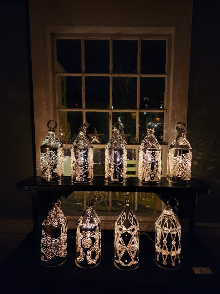 An array of ornate lanterns casting a warm, inviting glow on a shelf in a New England home, capturing the cozy and elegant ambiance of Christmas towns in the region. The intricate patterns on the lanterns create a beautiful play of light and shadow, contributing to the festive decor