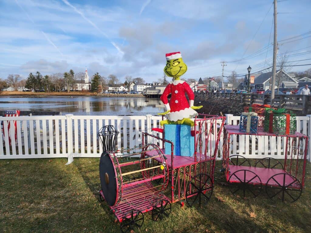 A whimsical outdoor Christmas display featuring a Grinch figure in a Santa outfit atop a pink train with wrapped presents, set against a scenic New England backdrop with a white church reflecting in the calm water