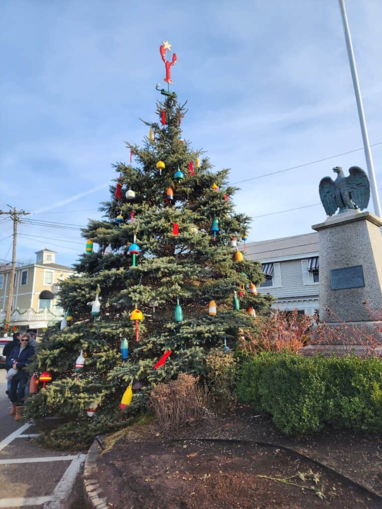 A festive outdoor Christmas tree stands in a town square in Kennebunkport Maine, uniquely decorated with an array of colorful fishing buoys and topped with a lobster ornament. The tree, set against a clear sky, brings a coastal twist to traditional holiday decorations, reflecting the local culture and heritage.