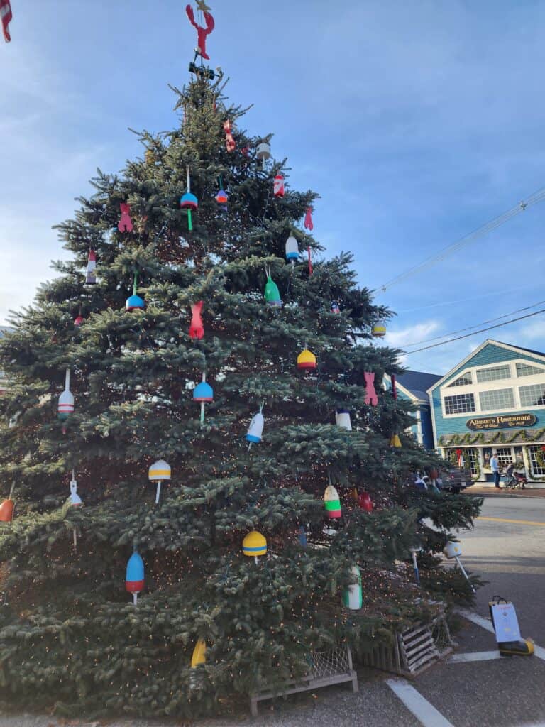 A large outdoor Christmas tree decorated with colorful lobster buoys and a red lobster topper, situated on a sunny day in front of Alison's Restaurant in Kennebunkport Maine, showcasing a festive seaside holiday tradition