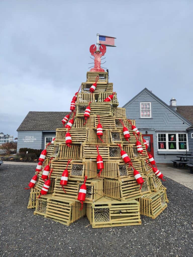 A festive lobster trap Christmas tree adorned with red and white buoys and an American flag, topped with a red lobster, in front of Fox's Lobster House, embodying the holiday spirit in a coastal New England setting