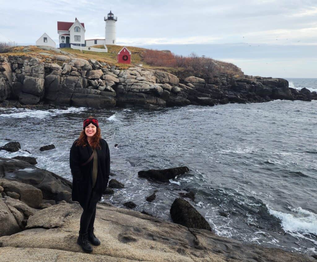 A woman in a red headband and black coat smiles at the camera, standing on rocky terrain with a picturesque New England lighthouse and white keeper's house in the background, epitomizing coastal charm