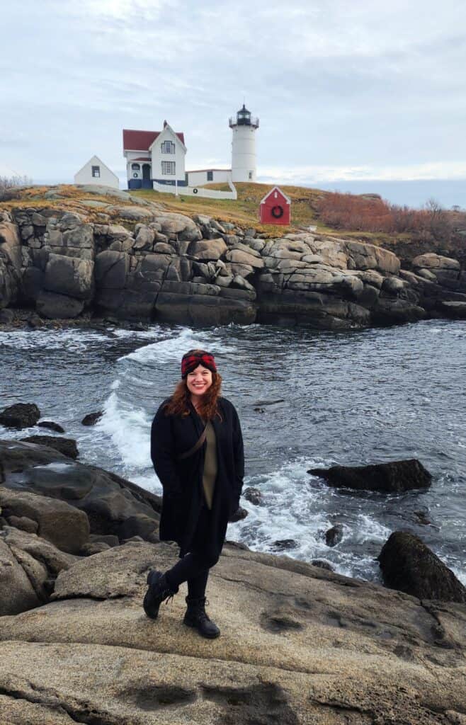 A woman stands on a rocky ledge in front of the Nubble Light in Maine.