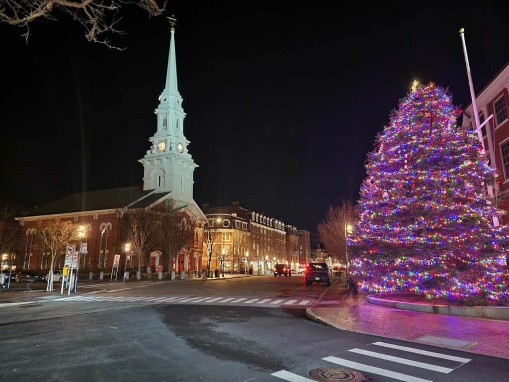 A picturesque New England town during Christmas, showcasing a vibrant Christmas tree adorned with colorful lights, standing beside a historic church with a tall white steeple. The scene is captured at night, emphasizing the festive glow against the quaint backdrop of classic architecture and quiet streets