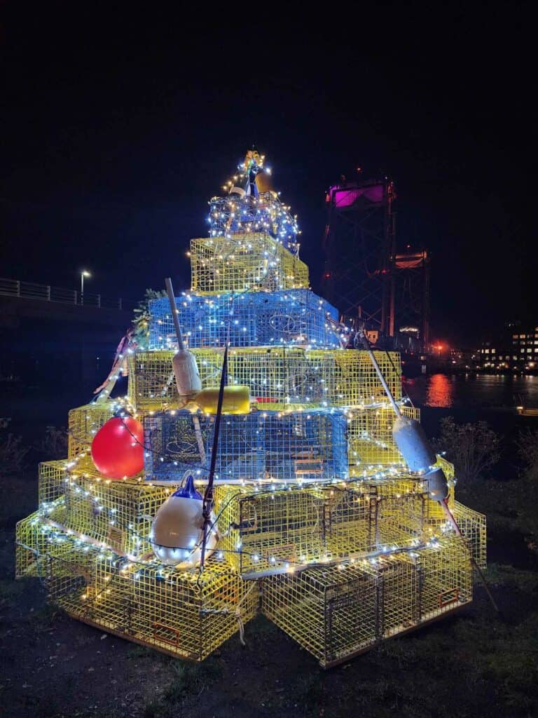 A night view of an innovative Christmas tree constructed from lobster traps, illuminated with string lights and adorned with large colorful buoys, with a drawbridge in the background, showcasing a unique New England holiday tradition