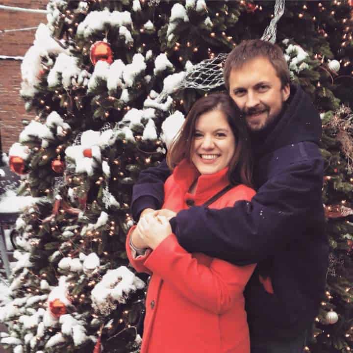 A couple poses in an embrace in front of a Christmas tree frosted with snow