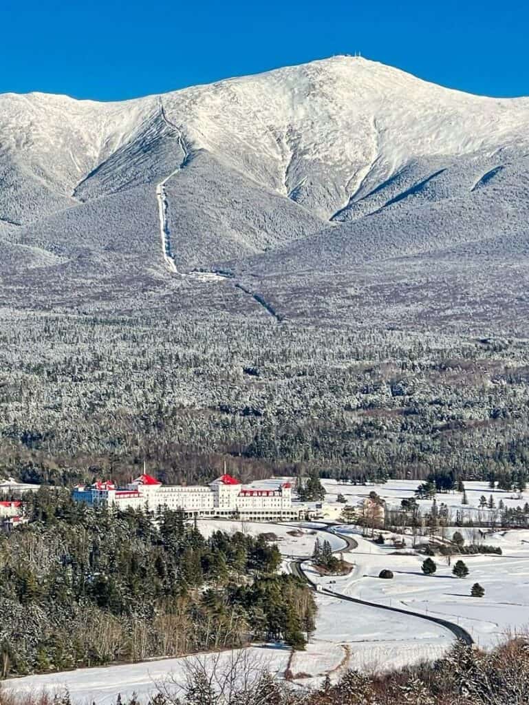 Snow-covered Mount Washington towers over the historic Omni Mount Washington Resort, a popular destination for winter getaways in New England, with a serpentine road leading up to the grand hotel amidst a forested landscape.